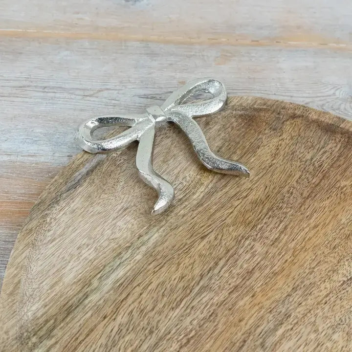 A silver metal bow-shaped decorative object resting on a round wooden board over a light wood surface.