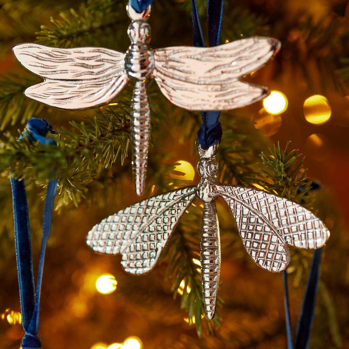 Silver dragonfly Christmas tree ornaments hanging from a green Christmas tree with warm lights in the background.