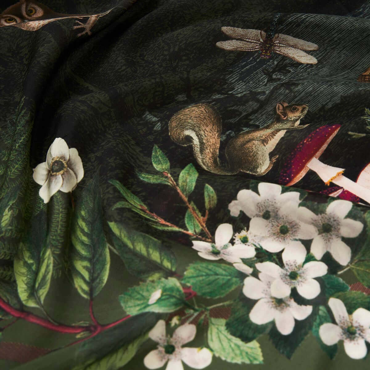 A close-up of a dark forest-themed scarf featuring a squirrel, dragonflies, white blossoms, green leaves, and red accents on a black and olive background.