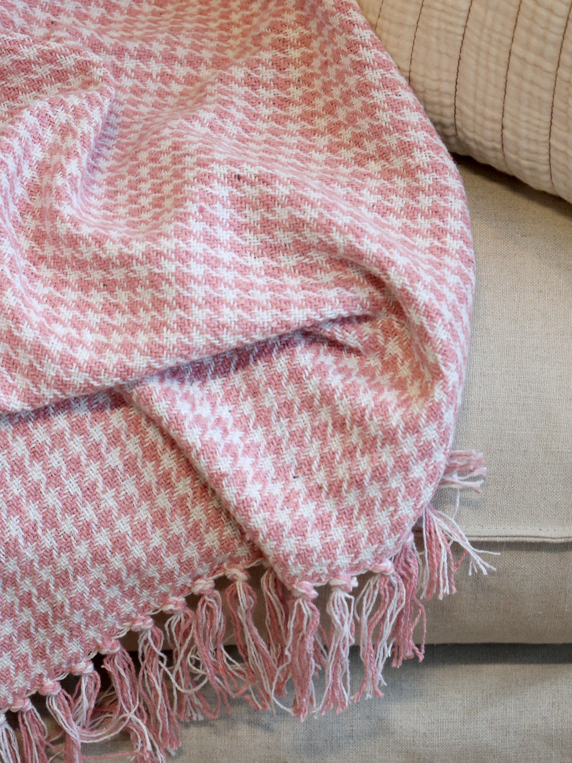 Pink and white checkered throw blanket with fringes draped over a beige sofa.
