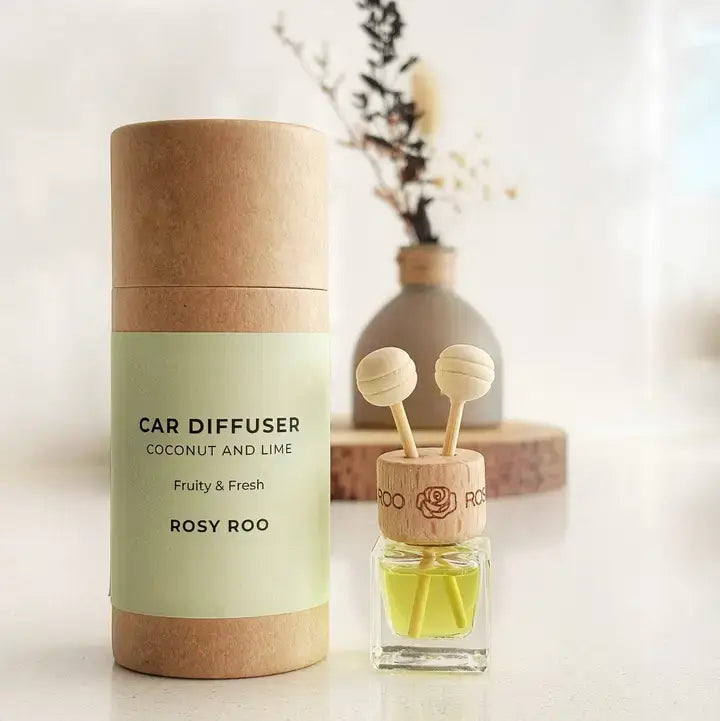A car diffuser set by Rosy Roo, featuring a kraft paper cylindrical packaging container with a pale green label next to the actual diffuser bottle. The diffuser has a wooden top with two hanging reeds and a yellow-green liquid inside, set against a softly blurred background with a vase of dried flowers.