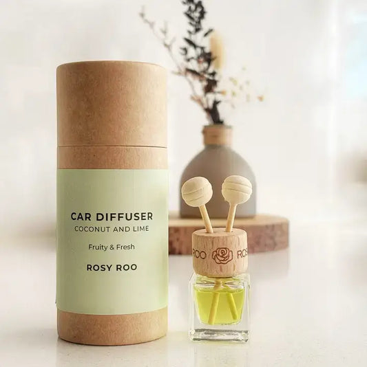 A car diffuser set by Rosy Roo, featuring a kraft paper cylindrical packaging container with a pale green label next to the actual diffuser bottle. The diffuser has a wooden top with two hanging reeds and a yellow-green liquid inside, set against a softly blurred background with a vase of dried flowers.