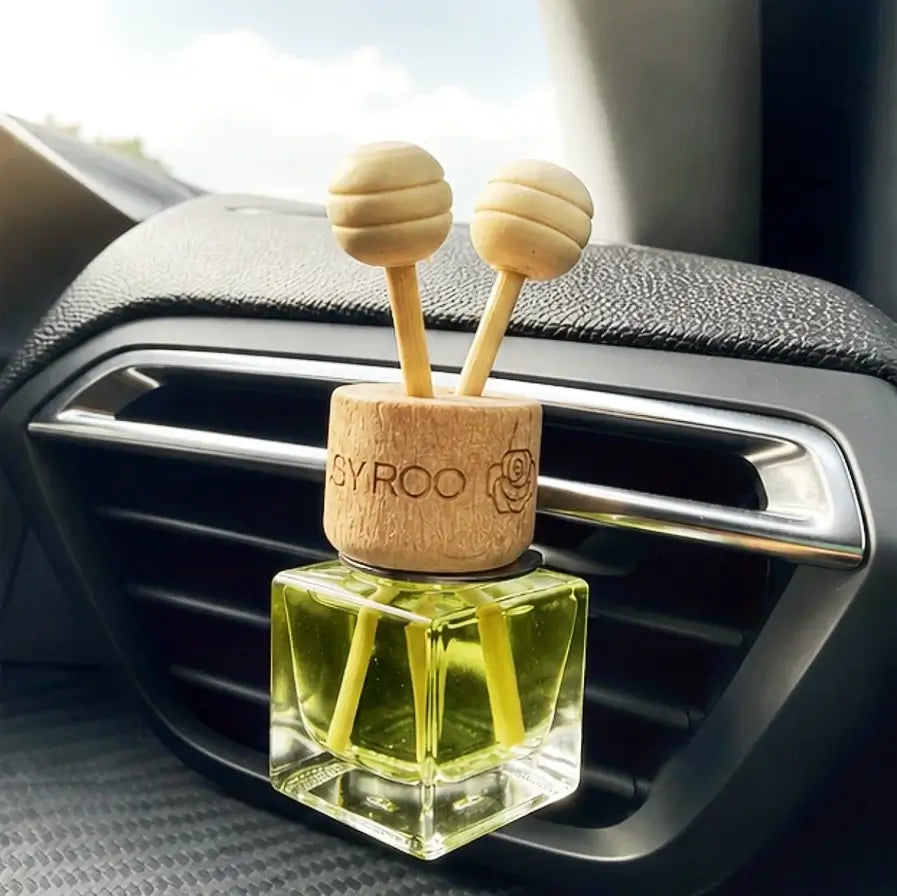 A car air freshener diffuser with a clear glass bottle filled with yellow-green liquid, a light wood cap engraved with 'SYROO', and two wooden honey dipper sticks, shown inserted into a car air vent.