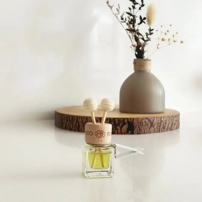 A small glass reed diffuser bottle with pale yellow liquid and two wooden diffuser sticks in a cork top, placed on a white surface. A round wood slice tray holds a matte taupe vase with dried stems in the background against a soft white wall.
