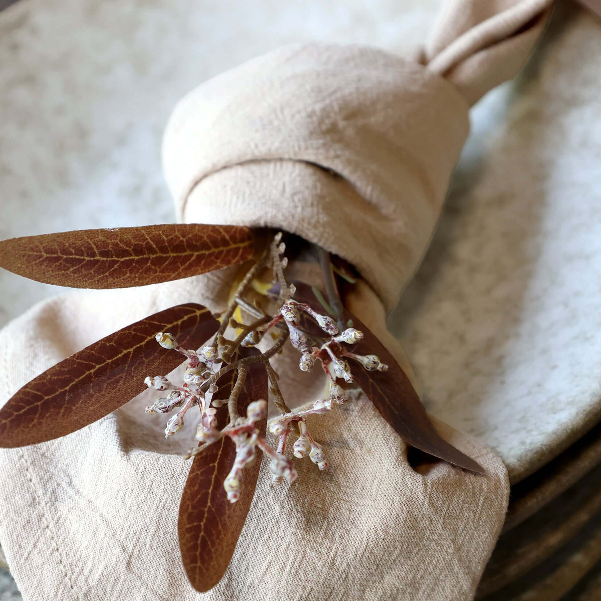 A close-up of a beige linen napkin tied in a knot with a dried brown leafy sprig, set against a soft neutral fabric background.