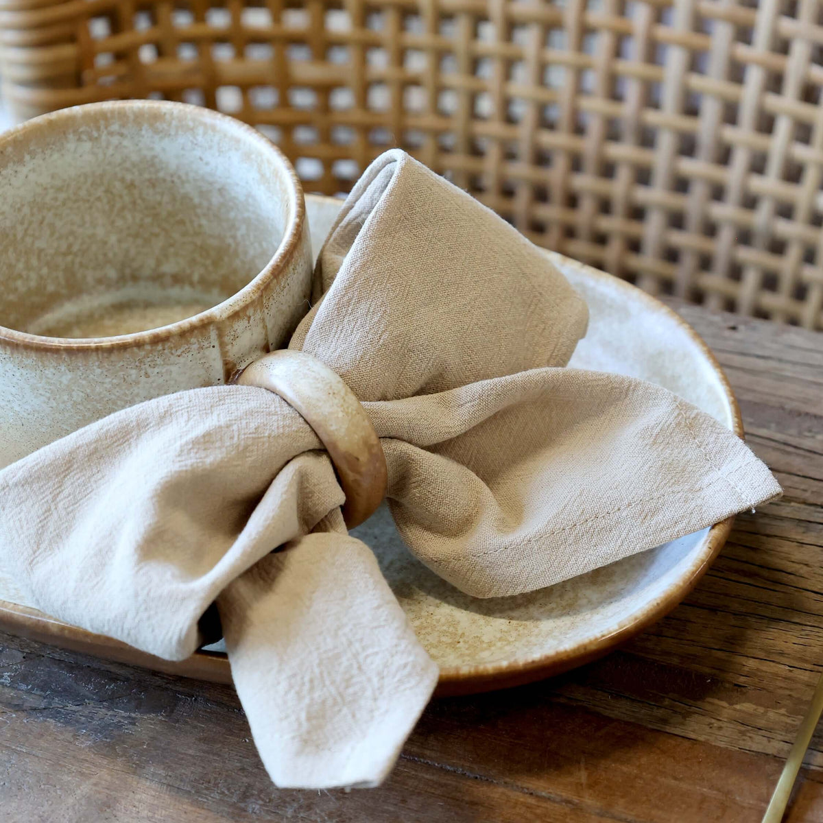 A beige linen napkin tied in a bow with a wooden ring, arranged on a ceramic plate next to a matching cup on a wooden table.