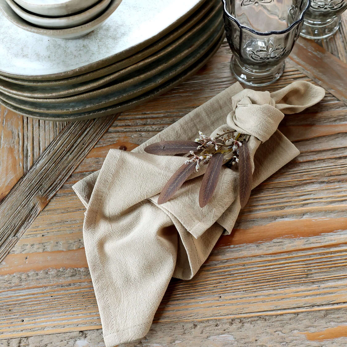 A beige linen napkin tied in a knot and decorated with dried brown leaves and small flowers, styled on a rustic wooden table with stacked plates and a glass in the background.