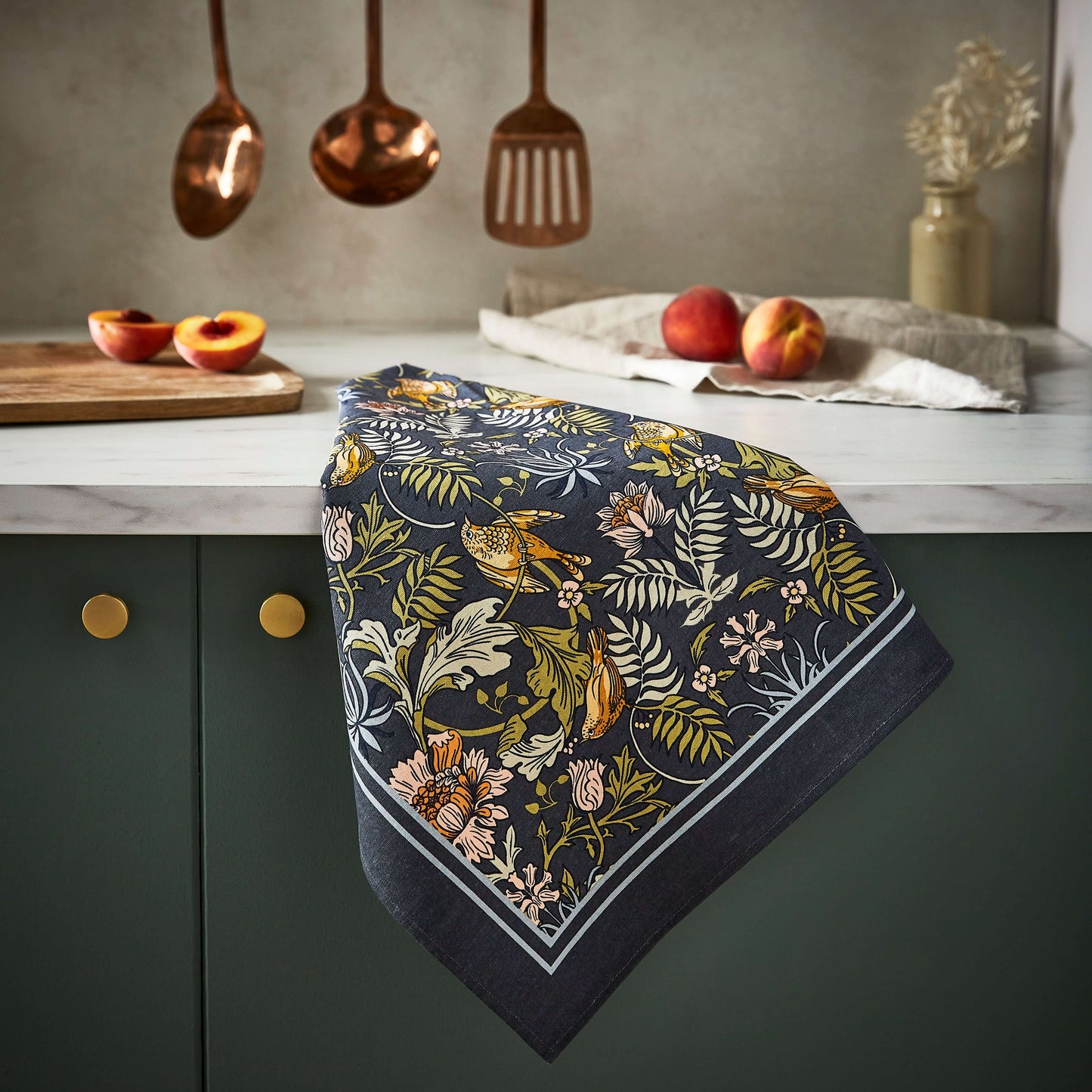 Floral patterned towel hanging on a kitchen cabinet with fruits and utensils in the background.