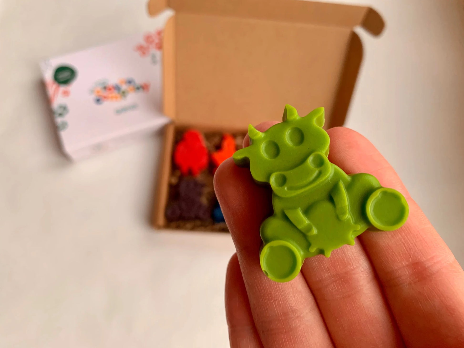 Hand holding a green cow-shaped crayon with a box of crayons in the background.
