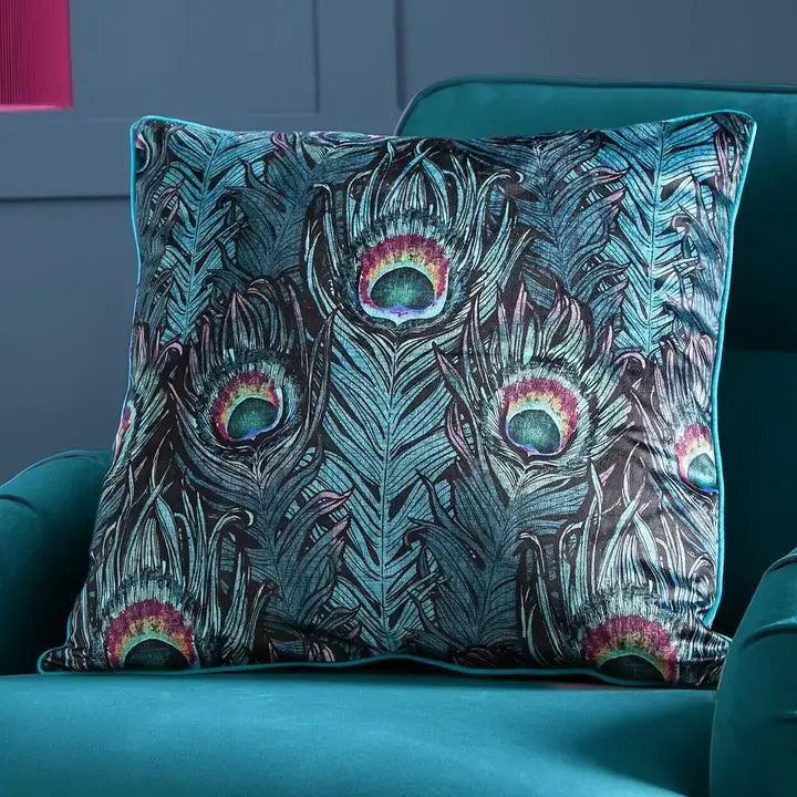 A square decorative throw pillow with a detailed peacock feather pattern in shades of teal, blue, black, and pink, styled on a teal velvet armchair.