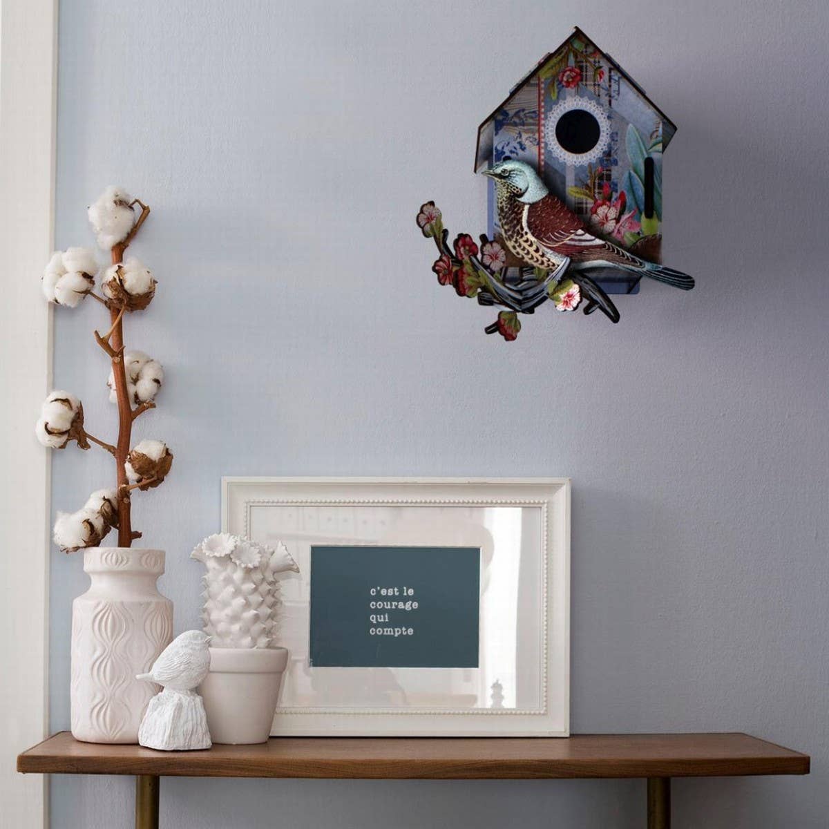 Decorative wall art with birds on a gray wall above a wooden shelf with decorative items.