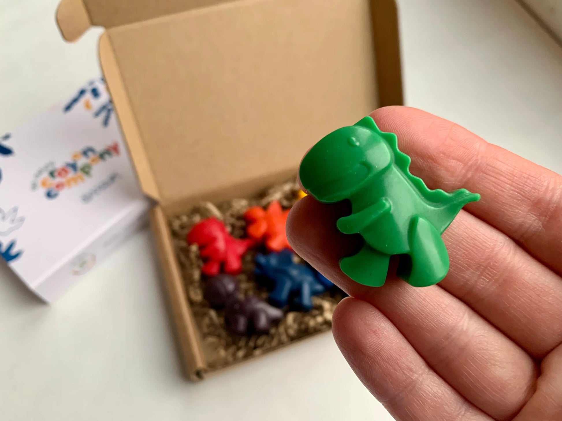 Green dinosaur-shaped crayon held by a hand with a box of crayons in the background.