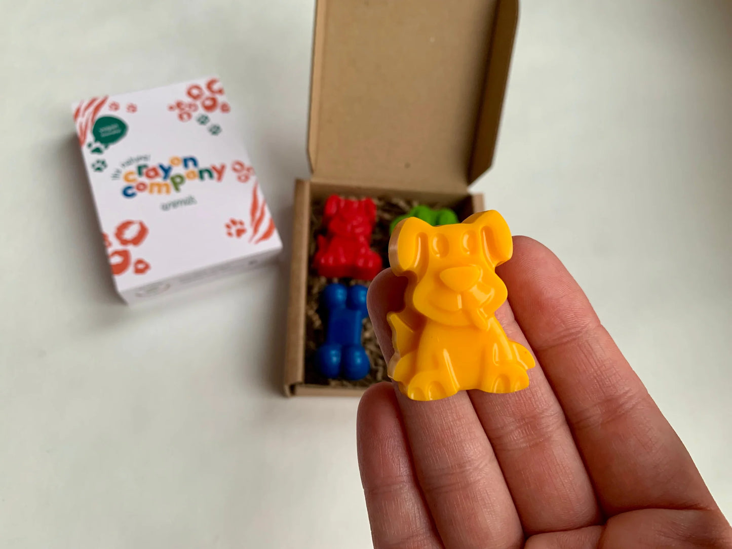 Colorful dog-shaped crayon held in a hand with a branded box in the background