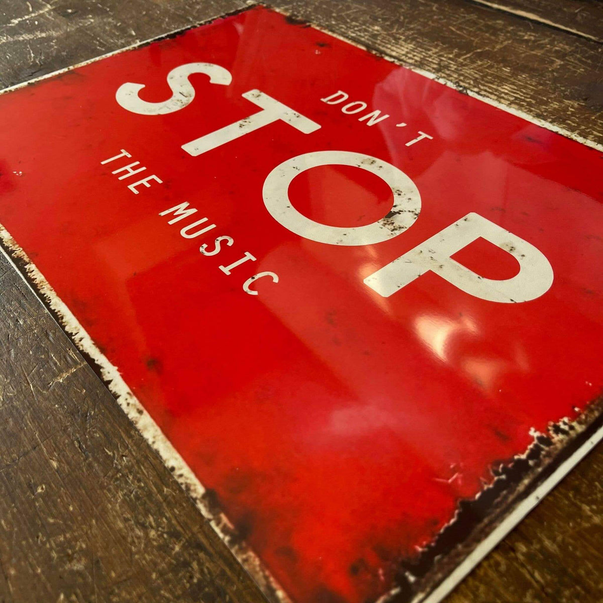 A distressed vintage-style red metal sign with white text reading 'DON'T STOP THE MUSIC,' photographed at an angle on a dark wooden surface.