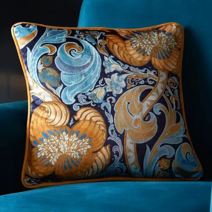 A square decorative throw pillow with an ornate floral and scroll pattern in gold, light blue, and white on a dark navy background, resting on a teal velvet armchair.