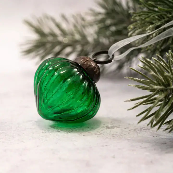A green ribbed glass Christmas ornament with a dark metal cap and a translucent ribbon loop, resting on a light surface with blurred pine branches in the background.