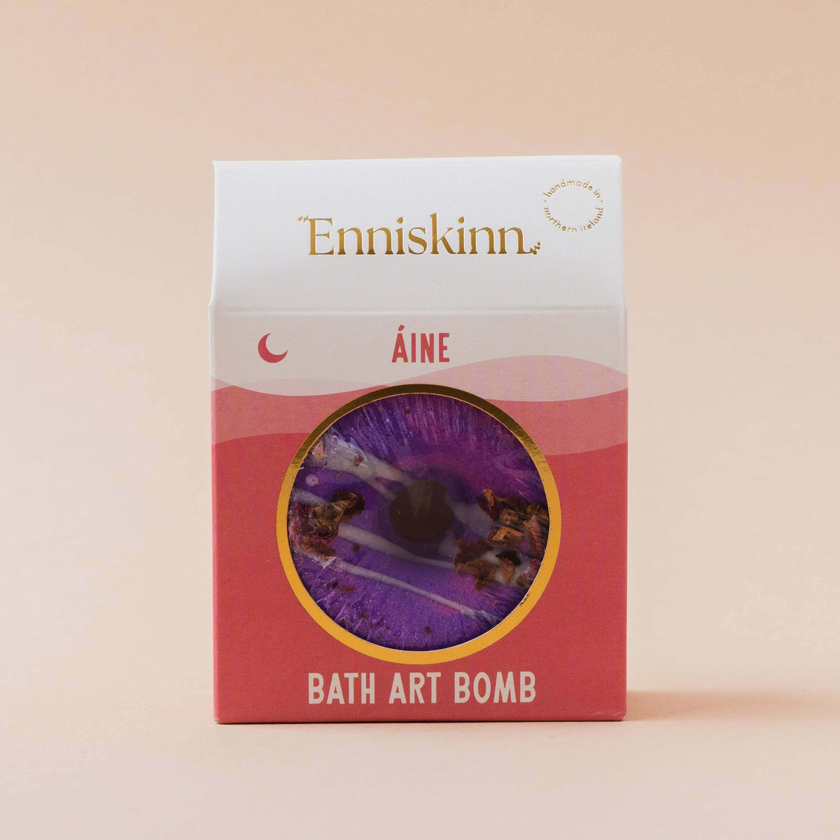 Enniskinn Éine Bath Art Bomb in a pink and white box with a circular window showing a purple bath bomb inside, set against a solid beige background.