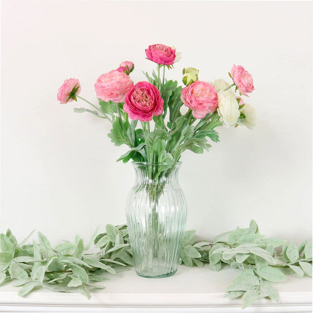 Clear glass vase with pink flowers on a white surface with green leaves.