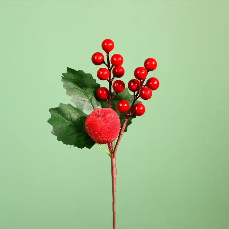 An artificial holly stem featuring glossy red berries, green leaves, and a textured red berry accent on a solid light green background.