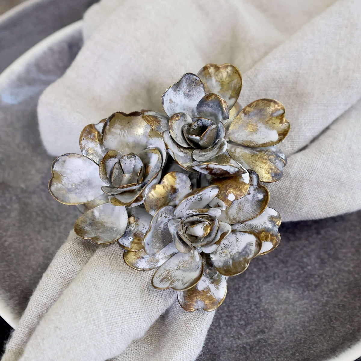 A close-up of a metallic floral napkin ring with silver and gold-toned petals, holding a light beige linen napkin on a dark gray plate.