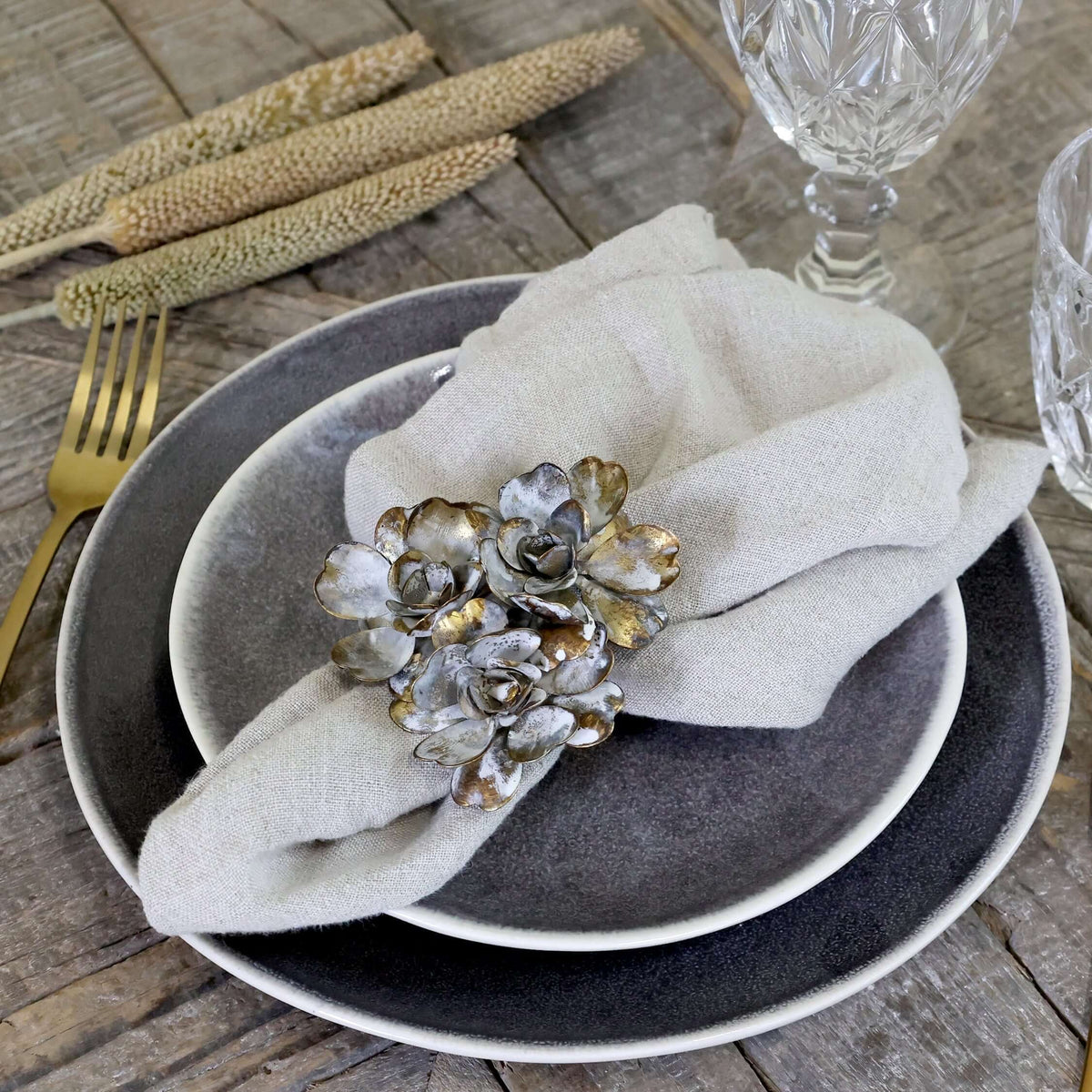 A styled table setting featuring a place setting with a gray and white layered plate, a light beige linen napkin, a metallic silver and gold flower napkin ring, gold-toned flatware, and clear cut-glass cups on a rustic wooden table.