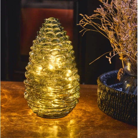 A pinecone-shaped glass table lamp glowing with warm yellow lights, placed on a reflective metallic surface next to a dark woven planter with dried branches.