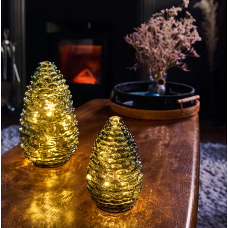 Two golden pine cone-shaped decorative lights with ribbed textured glass, glowing with warm yellow fairy lights, displayed on a wooden table in a cozy room setting with a fireplace in the background.