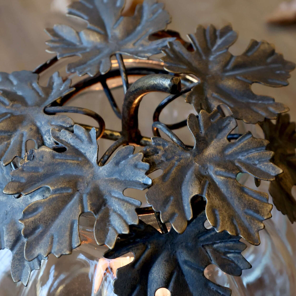 A close-up of a decorative metal grapevine wreath with textured dark gray leaves, draped over a clear ribbed glass pumpkin vase with a warm candle flame visible inside.