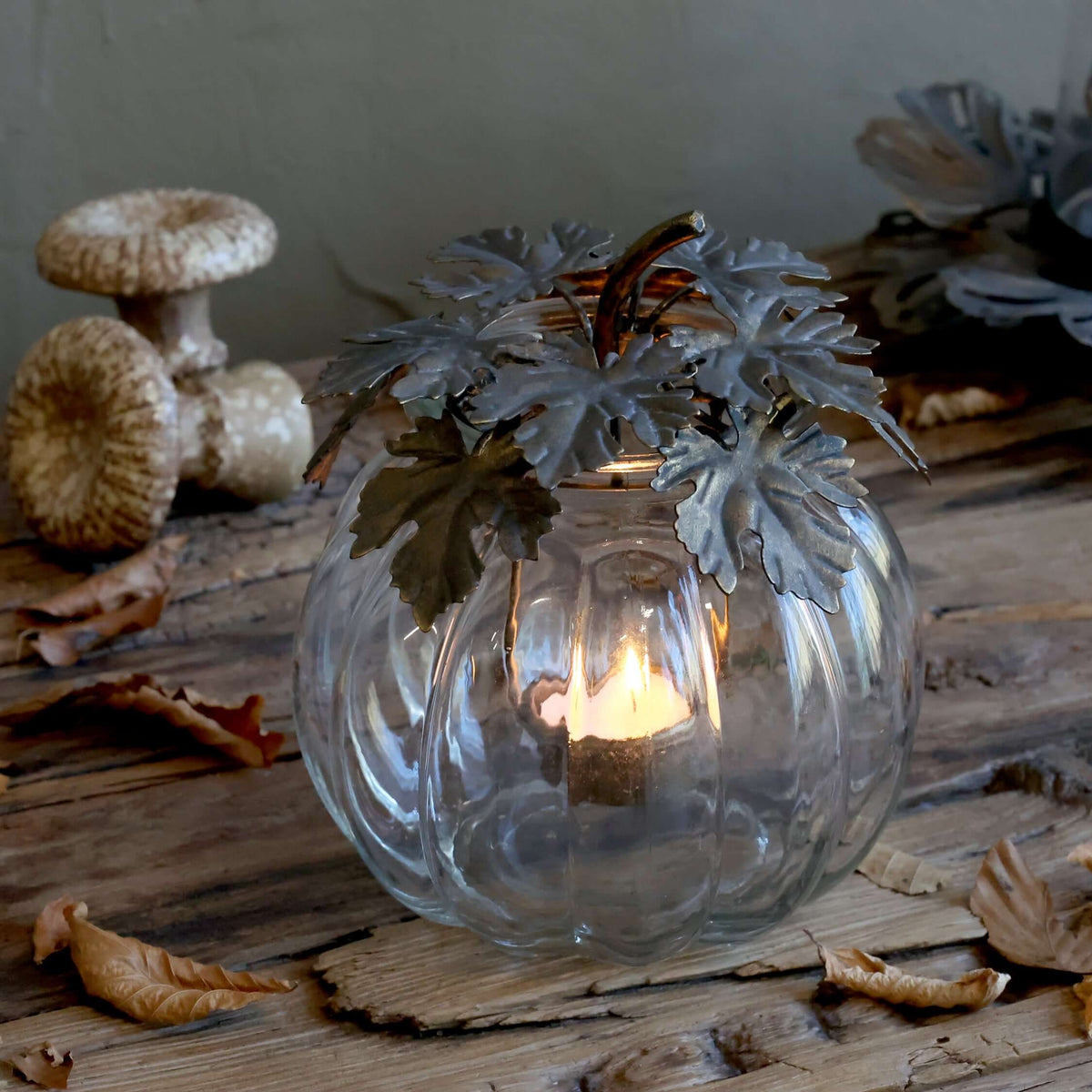 A clear ribbed glass pumpkin-shaped candle holder with a lit tea light inside, decorated with faux silver-gray leaves and a brown stem, set on a rustic wooden surface with dried leaves and mushrooms in the background.