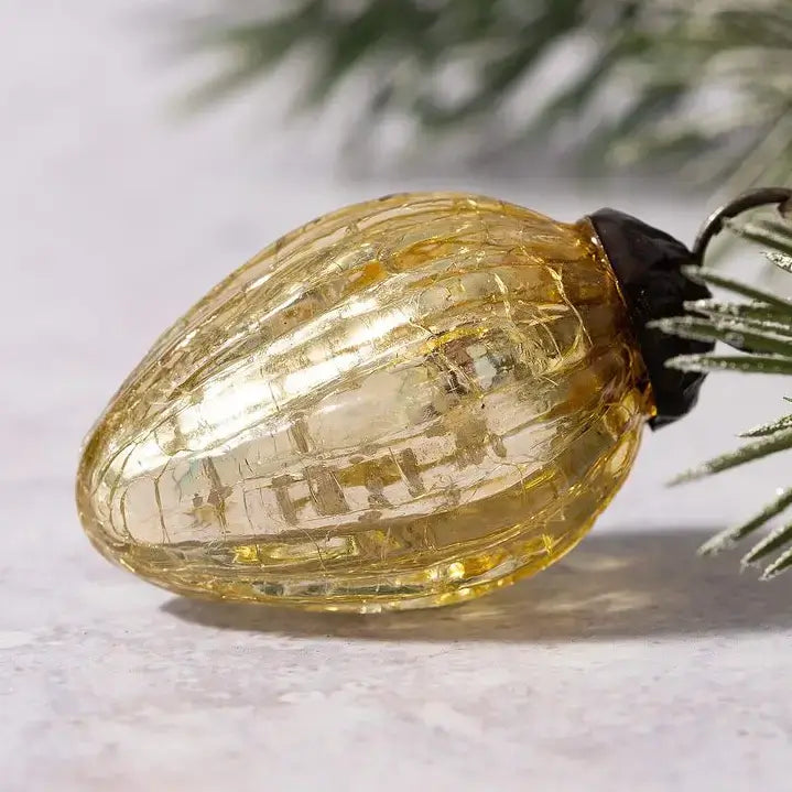 A single ribbed, translucent amber-yellow Christmas tree ornament with a black top, resting on a light textured surface with blurred pine needles in the background.