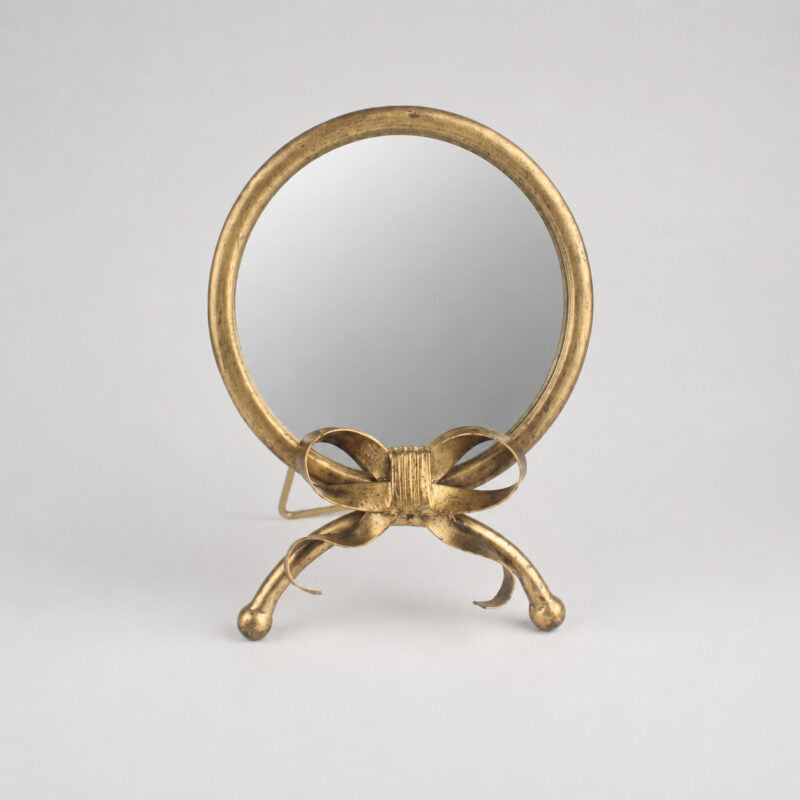 A small round tabletop mirror with a decorative gold-tone metal frame and bow-shaped stand, photographed against a plain light gray studio background.