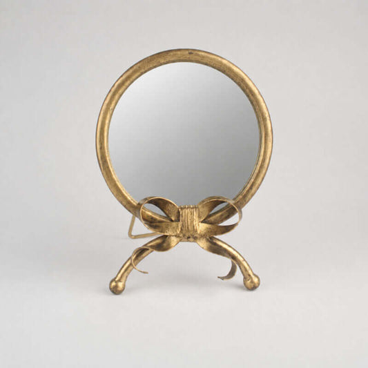 A small round tabletop mirror with a decorative gold-tone metal frame and bow-shaped stand, photographed against a plain light gray studio background.