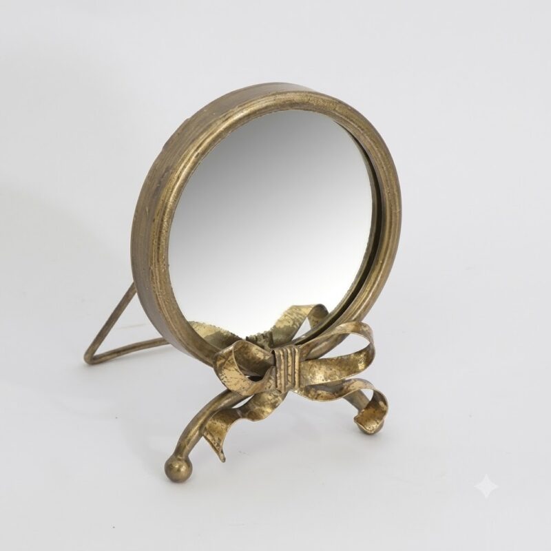 A small round tabletop mirror with a decorative antique gold metal frame and stand, featuring a bow detail at the base, set against a plain light gray background.