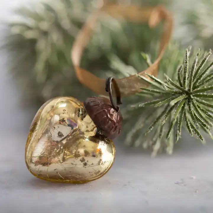 A heart-shaped glass ornament in a metallic gold finish with a small dark brown acorn detail and a tan ribbon loop, shown on a light surface with blurred green pine branches in the background.