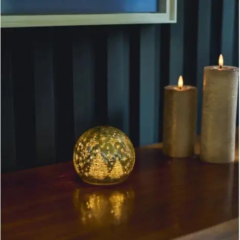 A gold glittery spherical LED lamo sits on a dark wooden dresser next to two lit pillar candles against a dark paneled wall.