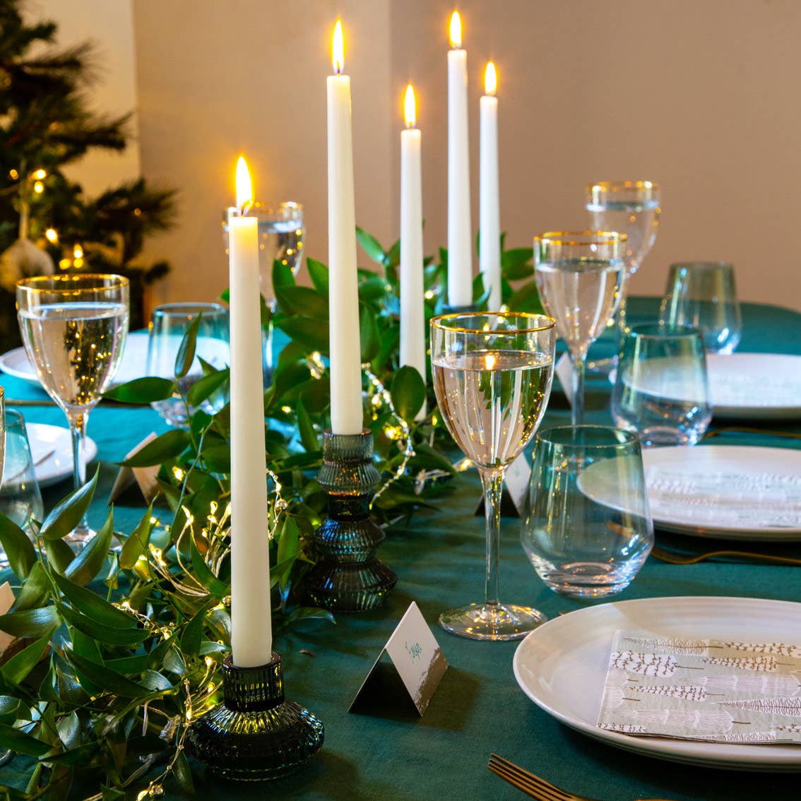 Elegant table setting with candles, glasses, and greenery on a teal tablecloth.
