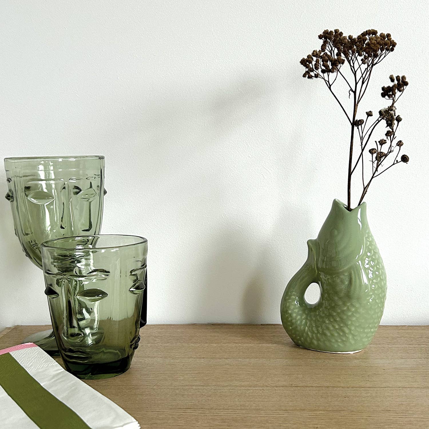 Two green glass face vases and a textured green ceramic vase with dried flowers on a wooden surface.