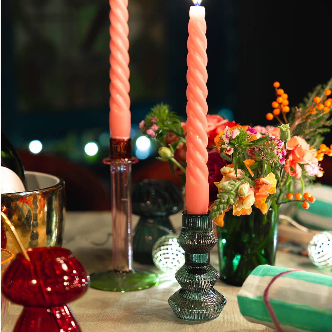 Twisted pink candles in decorative holders on a table with flowers and gift boxes.
