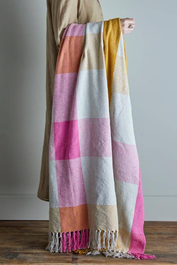 Person holding a colorful checkered blanket with tassels on a wooden floor.