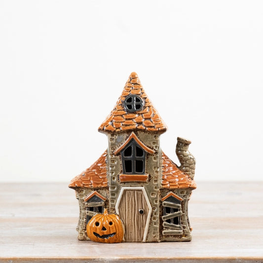 A small decorative haunted house ornament with an orange tiled roof, gray stone-textured walls, a wooden door, and a carved orange pumpkin in front, photographed on a light tabletop against a white background.