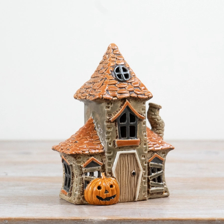 A small decorative haunted house figurine with orange tiled roofs, a carved jack-o'-lantern pumpkin at the base, and a gray stone chimney, photographed on a light wooden surface against a plain white background.