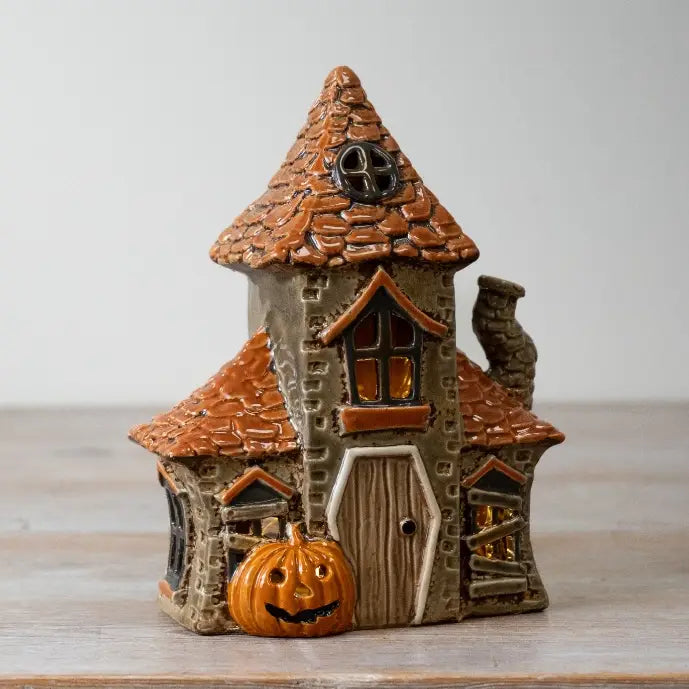 A small decorative haunted house figurine with an orange-brown tiled roof, gray stone-textured walls, a carved wooden door, and a jack-o'-lantern in front, photographed on a light tabletop against a soft neutral background.
