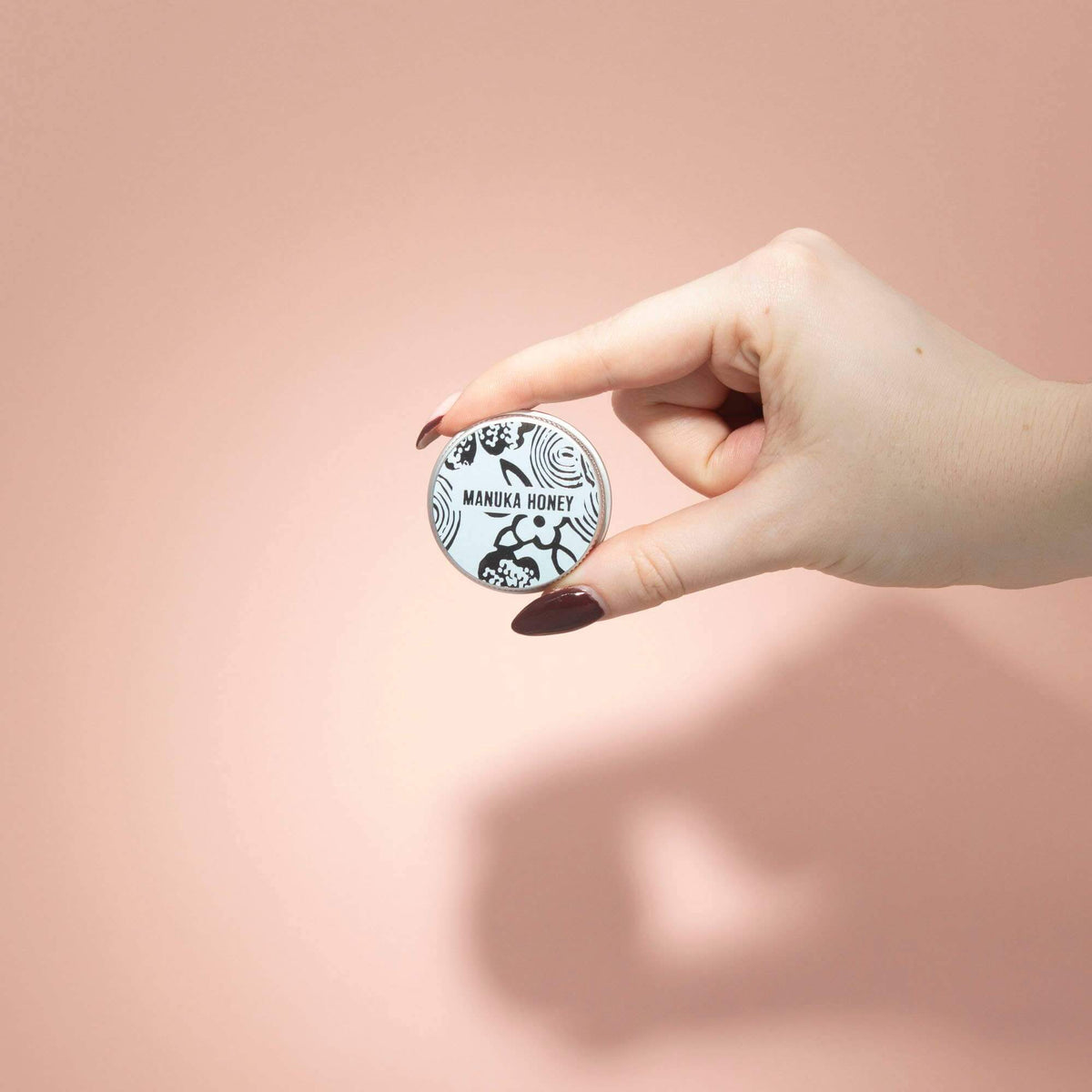  Hand holding a Manuka Honey lip balm tin with branding on a pink background
