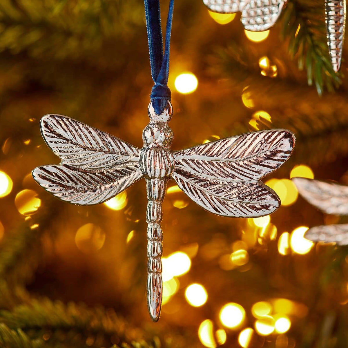 A silver dragonfly Christmas ornament hanging from a blue ribbon on a decorated Christmas tree with warm golden bokeh lights in the background.
