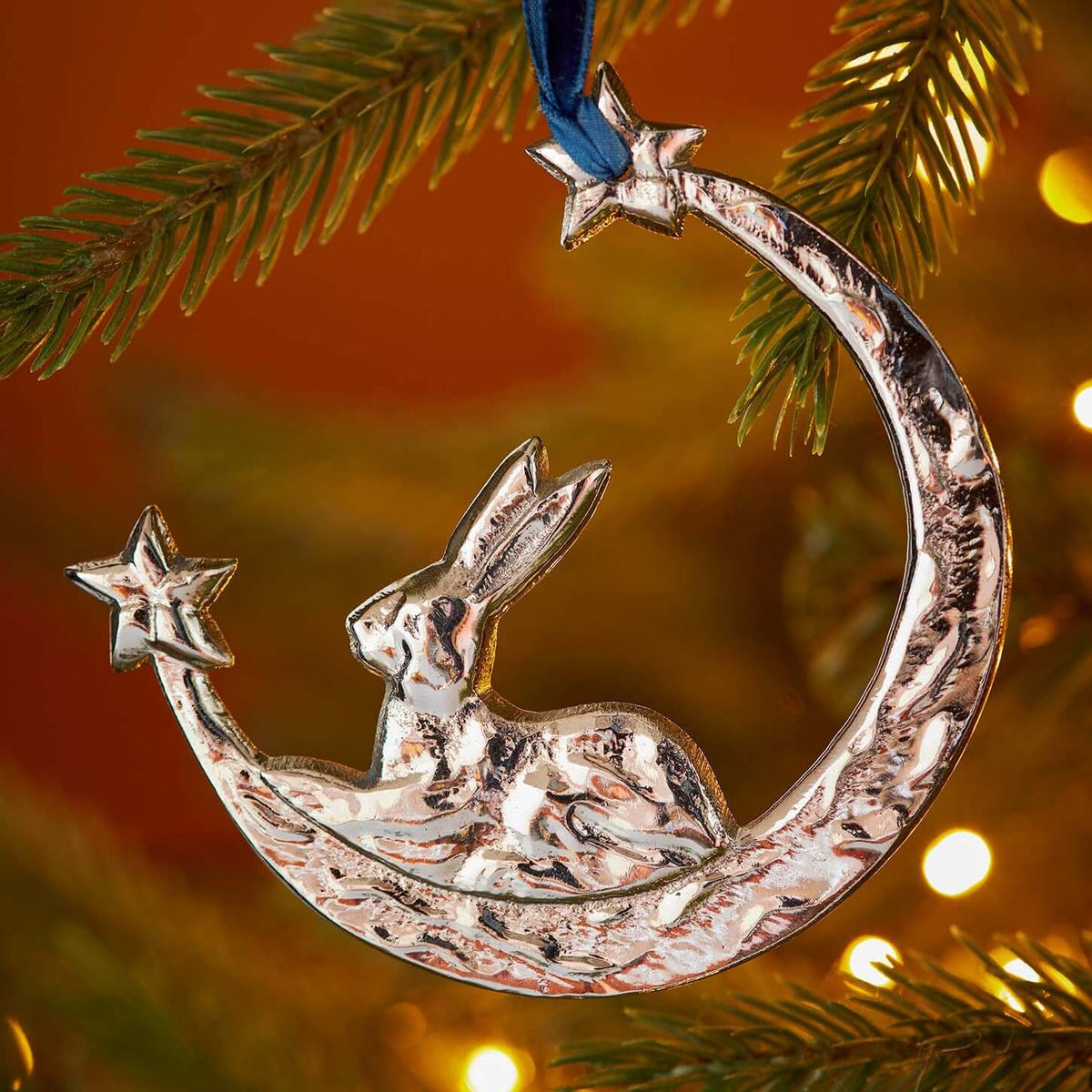 A silver-toned metal Christmas ornament shaped like a hare sitting on a crescent moon with two stars, hanging from a blue ribbon on a decorated Christmas tree branch with warm fairy lights.
