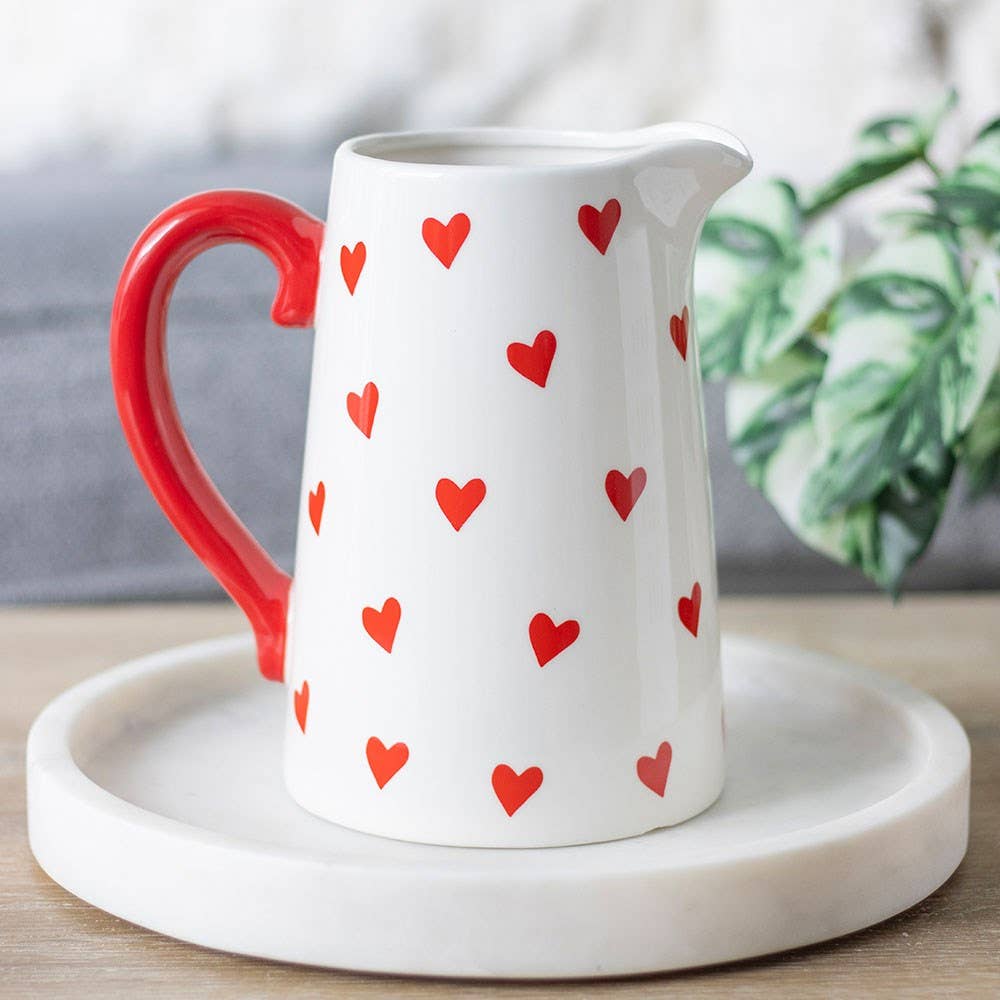 White pitcher with red heart patterns on a white plate, with a blurred plant in the background.