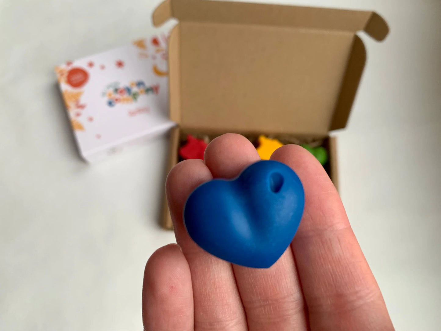 Blue heart-shaped crayon held in a hand with a box and colorful items in the background.