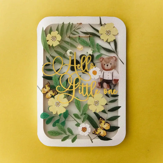 A greeting card featuring laser cut design with green leaves, yellow flowers, gold foil script reading 'Hello Little one', butterflies, and a small teddy bear wearing a shirt, set against a solid yellow background.