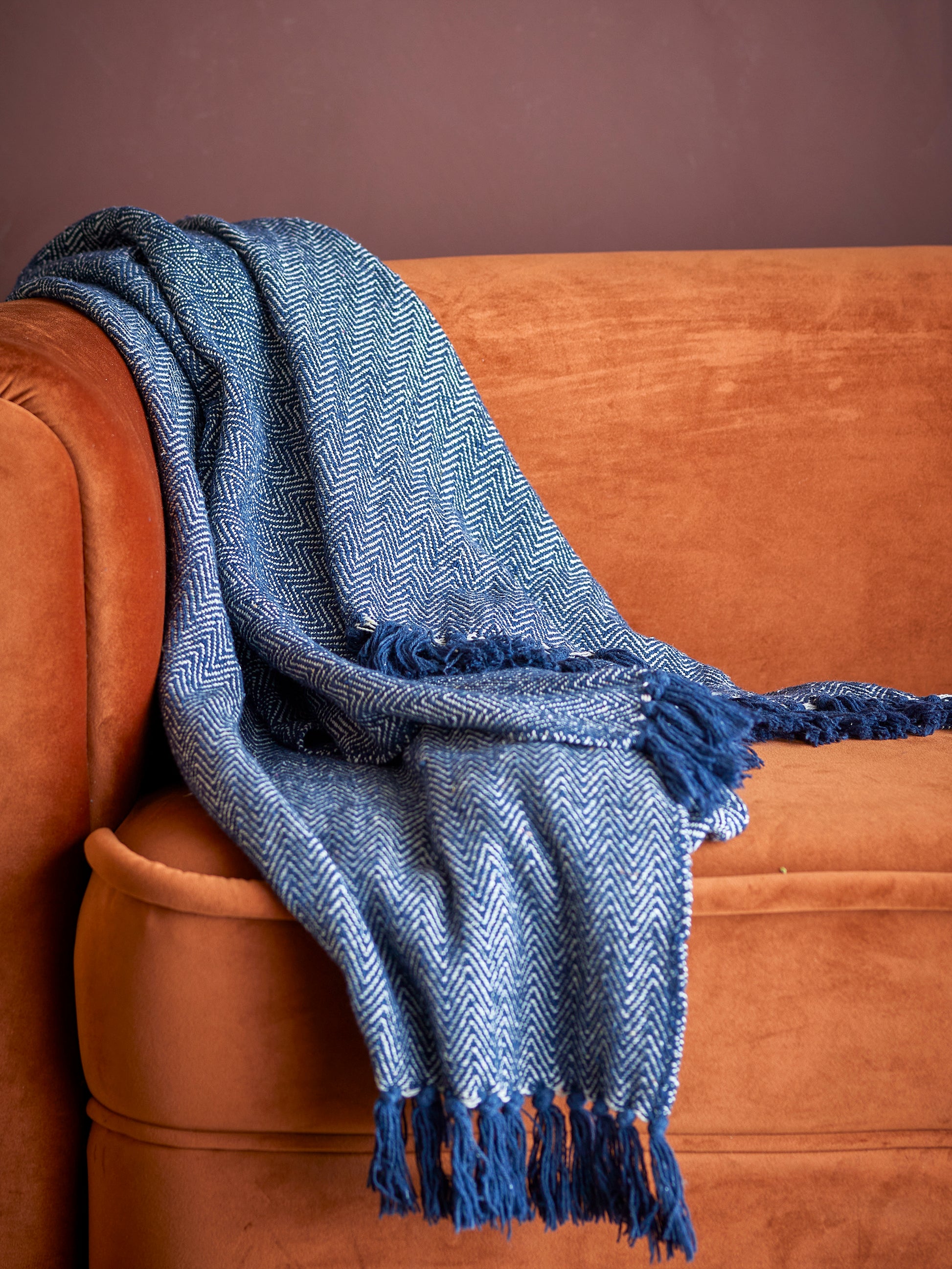 Blue woven blanket with tassels draped over a brown sofa against a brown background