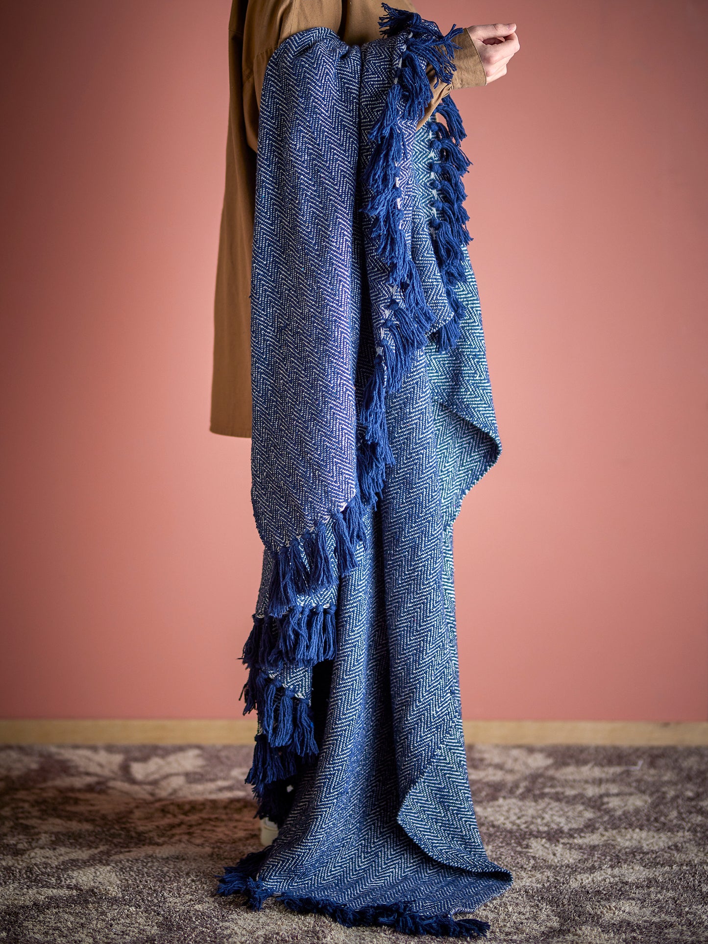 Person holding a blue knitted blanket with tassels against a pink wall.